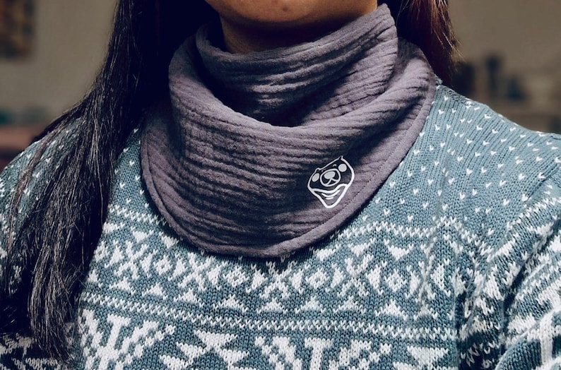 Puede incluir: Un pa&ntilde;uelo gris texturizado tipo babero con un logotipo de cara de oso de dibujos animados blanco. El pa&ntilde;uelo se lleva sobre un su&eacute;ter azul y blanco estampado. El pa&ntilde;uelo est&aacute; hecho de una tela suave y transpirable.
