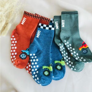 May include: Four pairs of children's socks in shades of orange, blue, and green. Each sock features a tractor graphic and the name "LIAM" in white. The soles have white star-shaped grips.