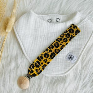 Puede incluir: Un babero de algodón blanco con dos broches de presión plateados y un clip para chupete de tela con estampado de leopardo con una cuenta de madera.