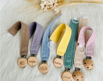 May include: A collection of baby pacifier clips in various colors, including brown, purple, blue, yellow, green, and pink. Each clip features a wooden disc with a name engraved on it. The clips are made of fabric and have a metal clasp.