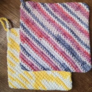May include: Two handmade crochet potholders. One is white and yellow, the other is white, pink, purple, and blue. Both have a diagonal striped pattern and a loop for hanging. The potholders are square.