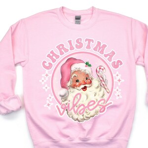 K&ouml;nnte beinhalten: Hellrosa Sweatshirt mit einem Vintage-Design von Santa Claus, der einen rosa Hut tr&auml;gt und eine Zuckerstange h&auml;lt. Die Worte "CHRISTMAS VIBES" sind in einem Kreis um Santa gedruckt.