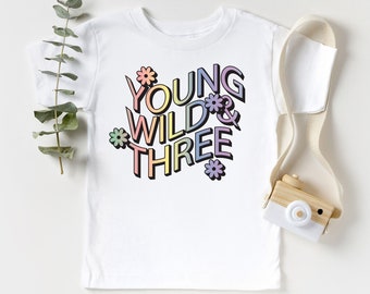May include: White toddler t-shirt with the text "YOUNG WILD & THREE" in colorful, outlined letters. The design includes flower accents. A wooden toy camera and a sprig of greenery are next to the shirt.