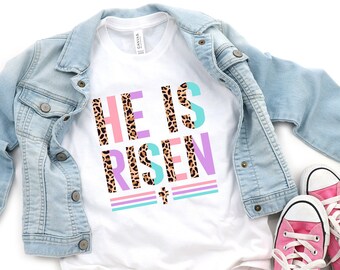 May include: A white t-shirt with the words "HE IS RISEN" in a colorful design, including leopard print, pink, teal, and purple. The shirt is paired with a light blue denim jacket and pink sneakers with white laces, creating a casual, spring-themed outfit.
