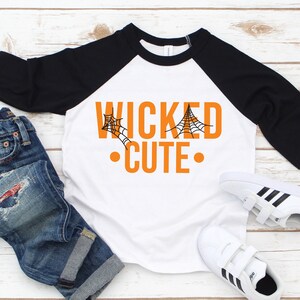 May include: A white and black raglan-style shirt with the words "WICKED CUTE" in orange, featuring spiderwebs. The shirt is paired with blue jeans and white sneakers with black stripes, perfect for Halloween.