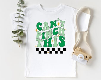 May include: White t-shirt with the text "CAN'T PINCH THIS" in green, with a retro font and a black and white checkered pattern at the bottom. The shirt has a crew neck and short sleeves. A sprig of greenery and a toy camera are next to the shirt.
