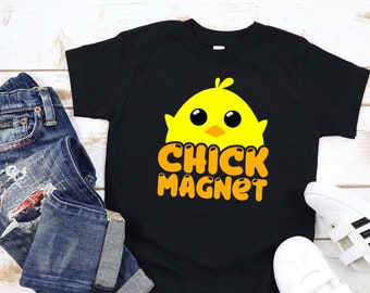 May include: Black t-shirt with a yellow chick graphic and the words "CHICK MAGNET" in orange. The shirt is paired with blue jeans and white sneakers with black stripes. A small potted plant is also visible.
