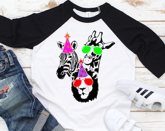 May include: A white and black raglan-style t-shirt featuring a graphic of a zebra, giraffe, and lion wearing party hats and sunglasses. The shirt is paired with blue jeans and white sneakers with black stripes.