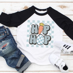 May include: White and black raglan style t-shirt with the words "HIP HOP" in a retro font, a carrot, and a checkered egg graphic. The shirt is paired with blue jeans and white sneakers with black stripes.