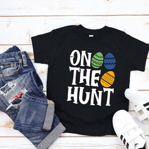 May include: Black t-shirt with the text "ON THE HUNT" and three colorful Easter egg designs. The shirt is paired with blue jeans and white sneakers with black stripes. A small potted plant is also visible.