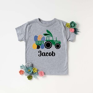 May include: A gray short-sleeved t-shirt featuring a green tractor pulling a wagon filled with colorful Easter eggs. The tractor has bunny ears. The name "Jacob" is printed in black script below the tractor design.