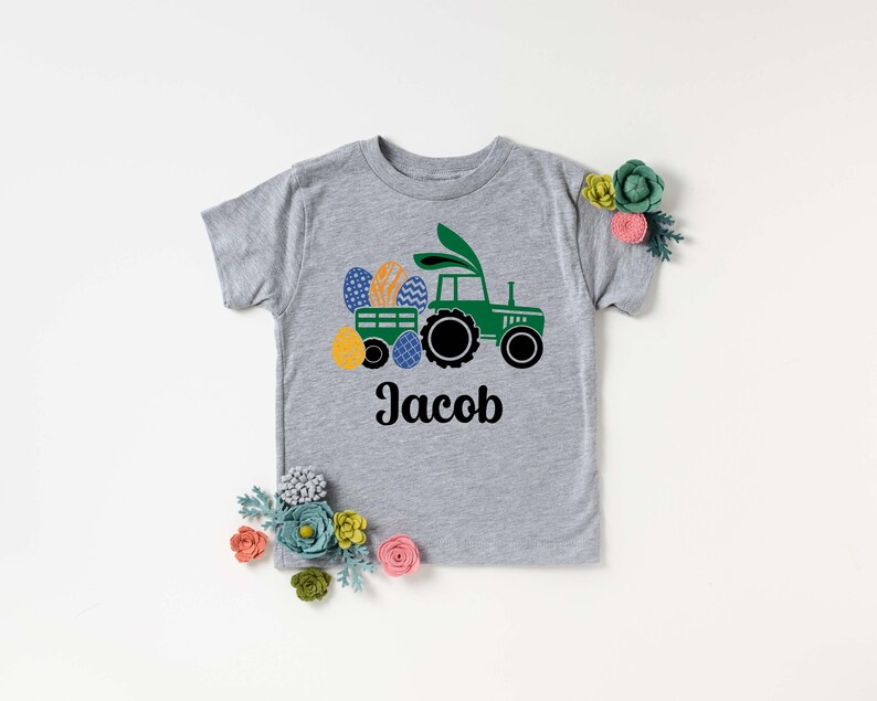 May include: A gray short-sleeved t-shirt featuring a green tractor pulling a wagon filled with colorful Easter eggs. The tractor has bunny ears. The name "Jacob" is printed in black script below the tractor design.