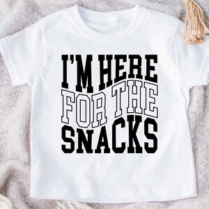 Puede incluir: Camiseta blanca con la frase "I'M HERE FOR THE SNACKS" en negrita y letras negras. El texto est&aacute; dispuesto en tres l&iacute;neas, ligeramente curvadas. La camiseta es de manga corta y cuello redondo.