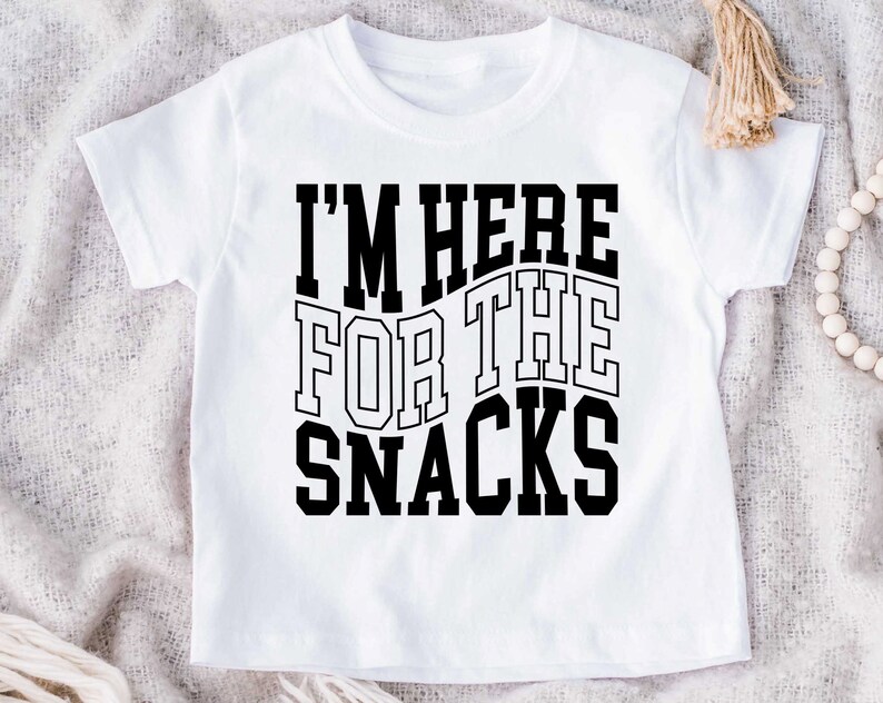 Puede incluir: Camiseta blanca con la frase "I'M HERE FOR THE SNACKS" en negrita y letras negras. El texto est&aacute; dispuesto en tres l&iacute;neas, ligeramente curvadas. La camiseta es de manga corta y cuello redondo.