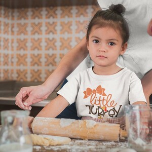 May include: A young child wearing a white t-shirt with the words "little TURKEY" in orange and brown. The child is in a kitchen, with a rolling pin and flour visible. An adult is nearby.