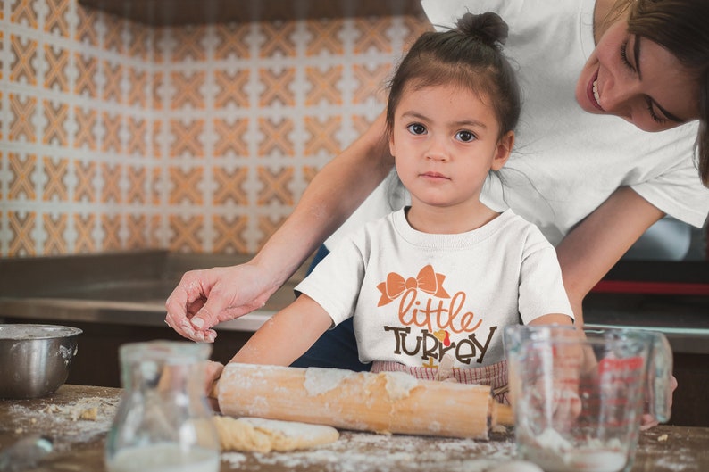 May include: A young child wearing a white t-shirt with the words "little TURKEY" in orange and brown. The child is in a kitchen, with a rolling pin and flour visible. An adult is nearby.