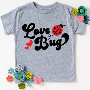 May include: A light gray t-shirt with the words "Love Bug" in black script and a red ladybug graphic. Two red hearts are to the left of the word "Bug". The shirt is decorated with felt flowers.