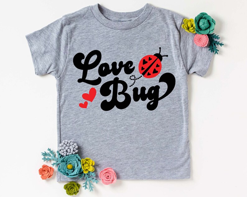 May include: A light gray t-shirt with the words "Love Bug" in black script and a red ladybug graphic. Two red hearts are to the left of the word "Bug". The shirt is decorated with felt flowers.