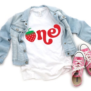 May include: A white t-shirt with a red strawberry and the word "one" in red script, paired with a light blue denim jacket and pink sneakers. The shirt is a casual style, perfect for a birthday celebration.
