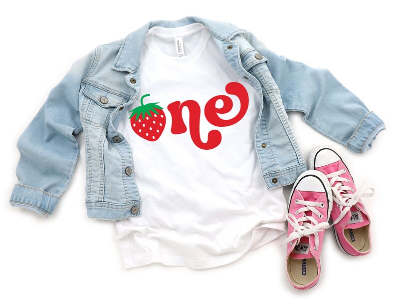 May include: A white t-shirt with a red strawberry and the word "one" in red script, paired with a light blue denim jacket and pink sneakers. The shirt is a casual style, perfect for a birthday celebration.
