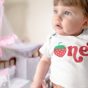 May include: White baby bodysuit with a red strawberry graphic and the word "one" in a playful font. The strawberry has green leaves and white seeds. The bodysuit is for a baby's first birthday.