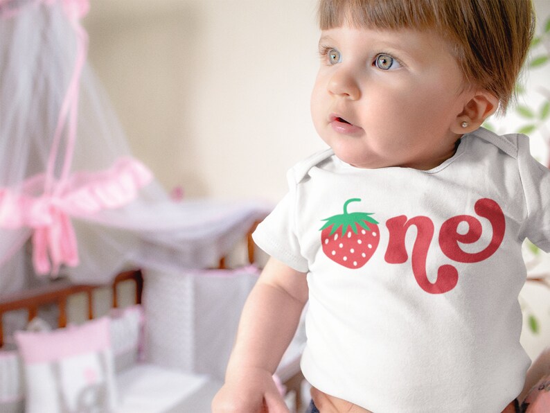 May include: White baby bodysuit with a red strawberry graphic and the word "one" in a playful font. The strawberry has green leaves and white seeds. The bodysuit is for a baby's first birthday.