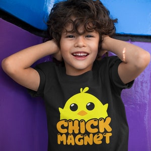 Puede incluir: Una camiseta negra con un dise&ntilde;o de pollito amarillo y el texto naranja "CHICK MAGNET". La camiseta la lleva un ni&ntilde;o sonriente con el pelo rizado casta&ntilde;o.