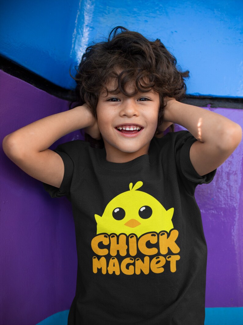 Puede incluir: Una camiseta negra con un dise&ntilde;o de pollito amarillo y el texto naranja "CHICK MAGNET". La camiseta la lleva un ni&ntilde;o sonriente con el pelo rizado casta&ntilde;o.