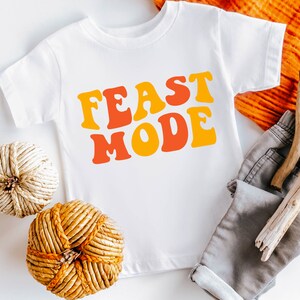 May include: White t-shirt with the words "FEAST MODE" in orange and yellow retro font. The shirt is displayed with gray shorts, decorative spheres, and wooden sticks. A festive, casual outfit.