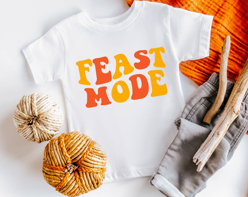 May include: White t-shirt with the words "FEAST MODE" in orange and yellow retro font. The shirt is displayed with gray shorts, decorative spheres, and wooden sticks. A festive, casual outfit.