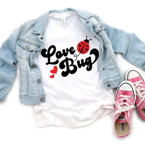 Peut inclure: Un t-shirt blanc avec l'inscription "Love Bug" en &eacute;criture noire et un dessin de coccinelle. Une veste en jean bleu clair et des baskets roses &agrave; lacets blancs sont &eacute;galement dans l'image. Deux c&oelig;urs rouges sont &agrave; c&ocirc;t&eacute; du mot "Bug".
