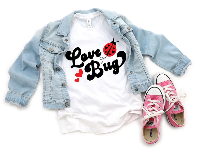 Peut inclure: Un t-shirt blanc avec l'inscription "Love Bug" en &eacute;criture noire et un dessin de coccinelle. Une veste en jean bleu clair et des baskets roses &agrave; lacets blancs sont &eacute;galement dans l'image. Deux c&oelig;urs rouges sont &agrave; c&ocirc;t&eacute; du mot "Bug".