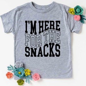 Puede incluir: Camiseta gris claro con el texto negro "I'M HERE FOR THE SNACKS". La camiseta est&aacute; adornada con flores de fieltro de colores.