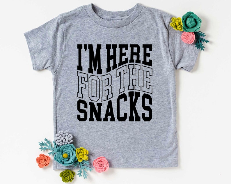 Puede incluir: Camiseta gris claro con el texto negro "I'M HERE FOR THE SNACKS". La camiseta est&aacute; adornada con flores de fieltro de colores.