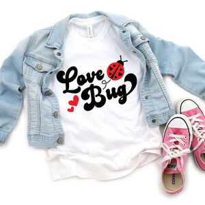May include: A white t-shirt with the words "Love Bug" in black script, a red ladybug graphic, and two red hearts. A light blue denim jacket and pink sneakers with white laces complete the ensemble.