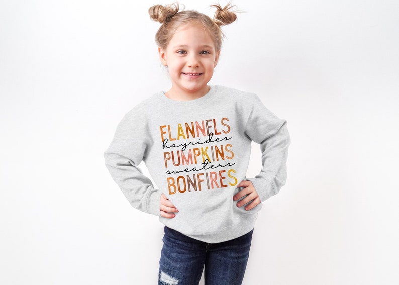Puede incluir: Una sudadera gris claro con las palabras "FLANNELS, hayrides, PUMPKINS, sweaters, BONFIRES" en una paleta de colores oto&ntilde;al. La sudadera la lleva una persona joven con las manos en las caderas.