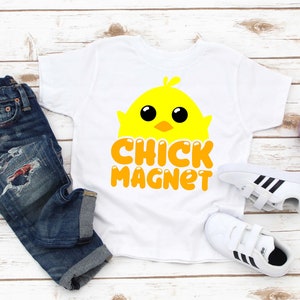 Puede incluir: Camiseta blanca con un dise&ntilde;o de pollito amarillo y las palabras "CHICK MAGNET" en naranja. La imagen tambi&eacute;n muestra vaqueros azules, zapatillas blancas con rayas negras y una peque&ntilde;a planta en maceta.