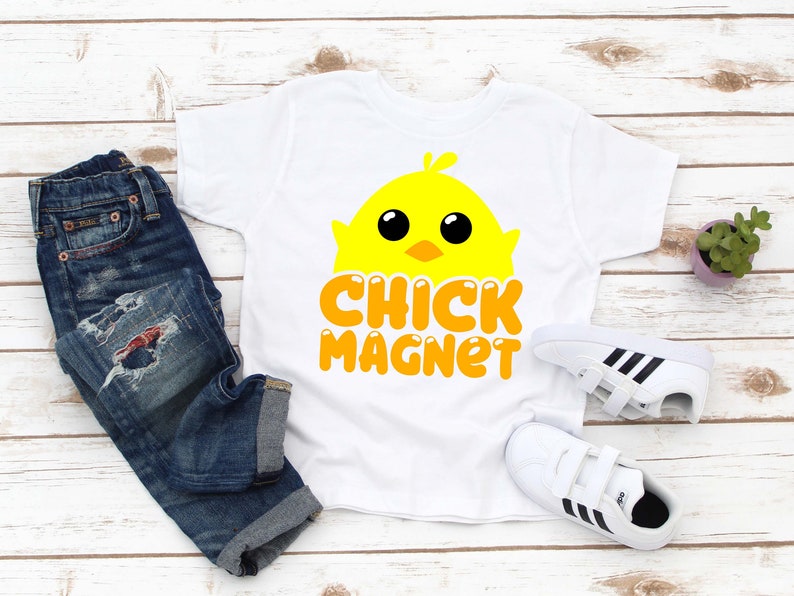 Puede incluir: Camiseta blanca con un dise&ntilde;o de pollito amarillo y las palabras "CHICK MAGNET" en naranja. La imagen tambi&eacute;n muestra vaqueros azules, zapatillas blancas con rayas negras y una peque&ntilde;a planta en maceta.