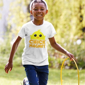Puede incluir: Un ni&ntilde;o con una camiseta blanca con un dibujo de pollito amarillo y las palabras "CHICK MAGNET" en naranja. Tambi&eacute;n lleva orejas de conejo y lleva una cesta colorida, corriendo al aire libre en un d&iacute;a soleado.