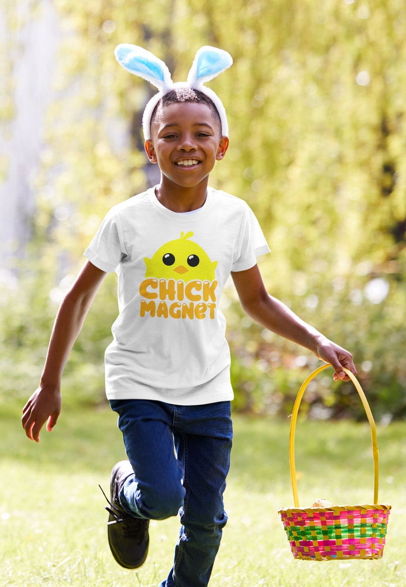 Puede incluir: Un ni&ntilde;o con una camiseta blanca con un dibujo de pollito amarillo y las palabras "CHICK MAGNET" en naranja. Tambi&eacute;n lleva orejas de conejo y lleva una cesta colorida, corriendo al aire libre en un d&iacute;a soleado.