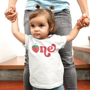 May include: A white t-shirt with a red strawberry graphic and the word "one" in red script. The shirt is worn by a young child. The child is being held by an adult.