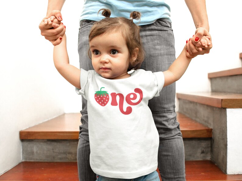 May include: A white t-shirt with a red strawberry graphic and the word "one" in red script. The shirt is worn by a young child. The child is being held by an adult.