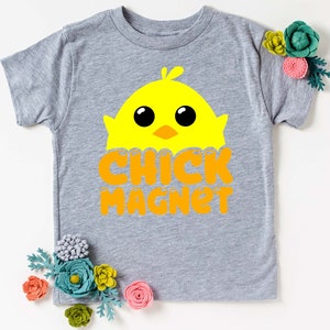 Puede incluir: Una camiseta gris de manga corta con un gr&aacute;fico de pollito amarillo y el texto naranja "CHICK MAGNET". El dise&ntilde;o est&aacute; centrado en la parte delantera de la camiseta. Flores decorativas est&aacute;n colocadas alrededor de la camiseta.