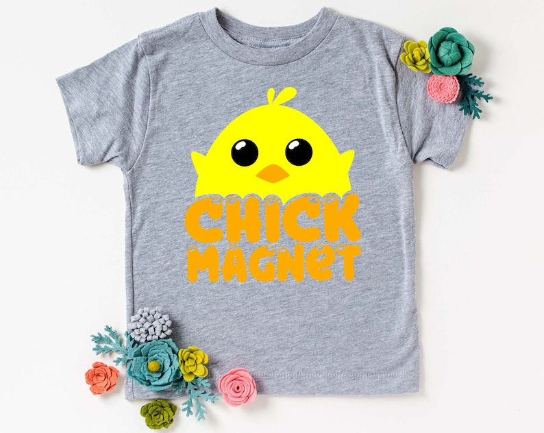Puede incluir: Una camiseta gris de manga corta con un gr&aacute;fico de pollito amarillo y el texto naranja "CHICK MAGNET". El dise&ntilde;o est&aacute; centrado en la parte delantera de la camiseta. Flores decorativas est&aacute;n colocadas alrededor de la camiseta.