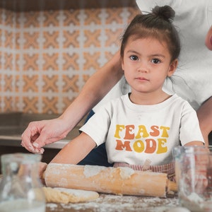 May include: A young child wearing a white t-shirt with the words "FEAST MODE" in orange and yellow lettering. The child is in a kitchen, with a rolling pin and dough visible. An adult is nearby.