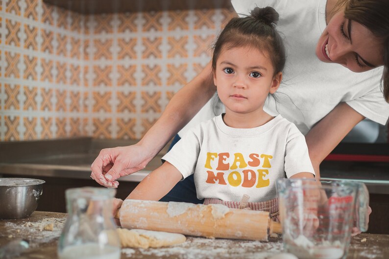 May include: A young child wearing a white t-shirt with the words "FEAST MODE" in orange and yellow lettering. The child is in a kitchen, with a rolling pin and dough visible. An adult is nearby.