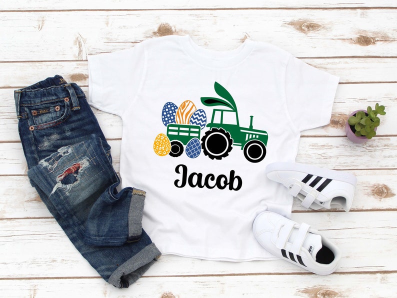 May include: White toddler t-shirt featuring a green tractor pulling a wagon filled with Easter eggs. The tractor has bunny ears. The name "Jacob" is printed in black below the tractor. A pair of blue jeans and white sneakers are next to the shirt.