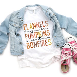 Puede incluir: Una camiseta blanca con las palabras "FLANNELS, hayrides, PUMPKINS, sweaters, BONFIRES" en colores oto&ntilde;ales. Una chaqueta vaquera azul claro est&aacute; sobre la camiseta, con zapatillas rosas con cordones blancos en la parte inferior.