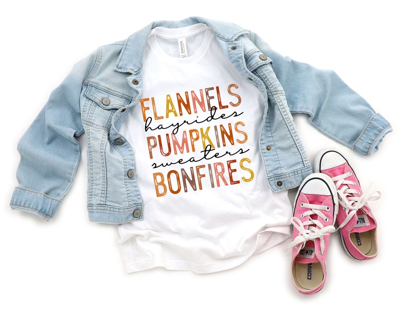 Puede incluir: Una camiseta blanca con las palabras "FLANNELS, hayrides, PUMPKINS, sweaters, BONFIRES" en colores oto&ntilde;ales. Una chaqueta vaquera azul claro est&aacute; sobre la camiseta, con zapatillas rosas con cordones blancos en la parte inferior.