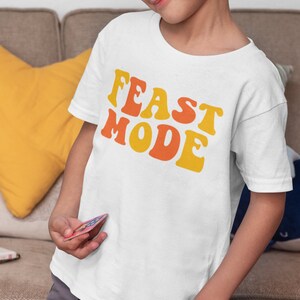 May include: White t-shirt with the words "FEAST MODE" in orange and yellow retro font. The shirt is worn by a child. The child is holding a card and is sitting at a wooden table with other cards.